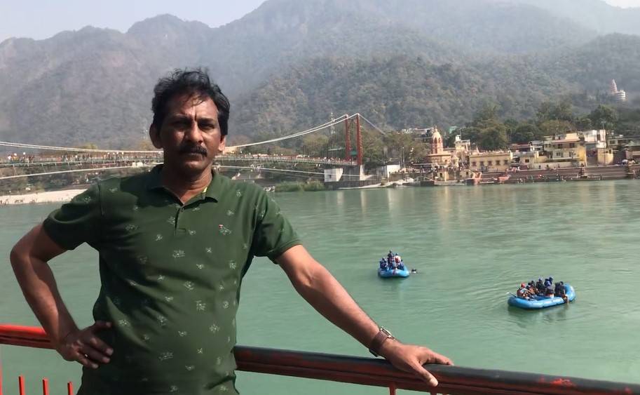 the author stands by a red railing overlooking the Ganges River in Rishikesh, with the iconic Laxman Jhula bridge, surrounding hills, and rafting boats visible in the background.