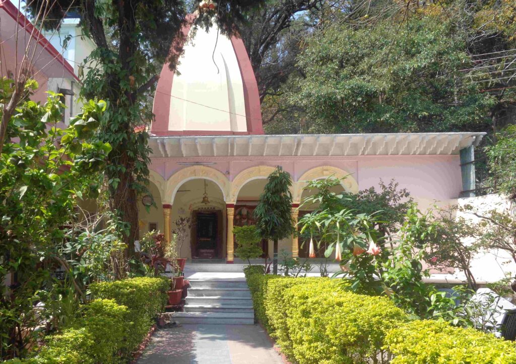 A serene temple in Rishikesh surrounded by lush greenery and flowering plants, with steps leading to the main entrance under arched verandas
