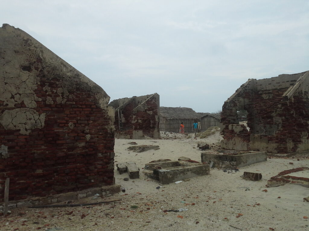 "Ruins of abandoned buildings in Dhanushkodi, Tamil Nadu, with weathered brick walls standing in a sandy coastal landscape and a few visitors in the distance