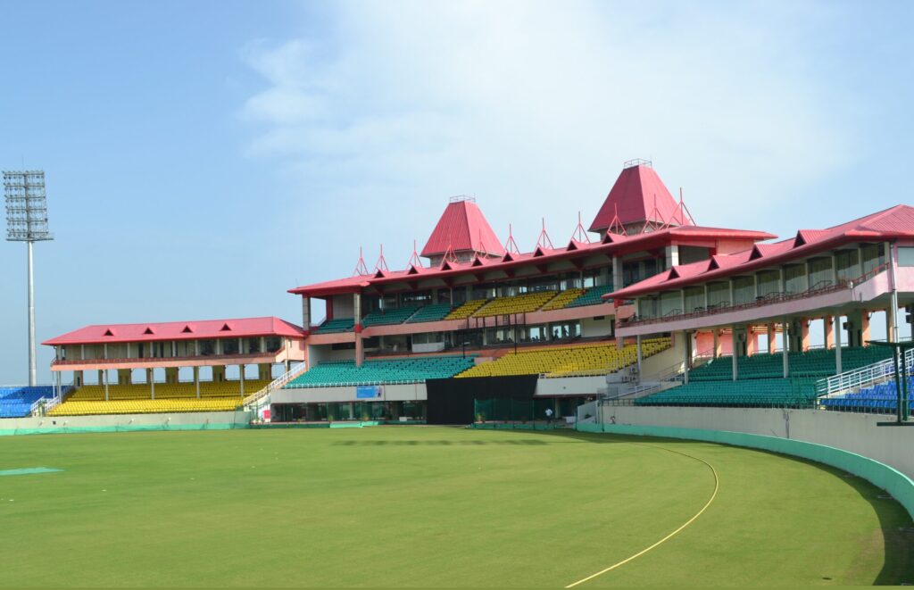 The colourful seating stands and Tibetan-style pavilion of the Dharamshala Cricket Stadium, with its vibrant red roofs and well-maintained green outfield under a clear sky.