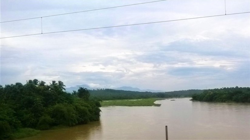 A scenic river landscape captured through a train window, showing a wide muddy river winding between lush green banks, with distant hills and a cloudy sky creating a calm travel-story moment.