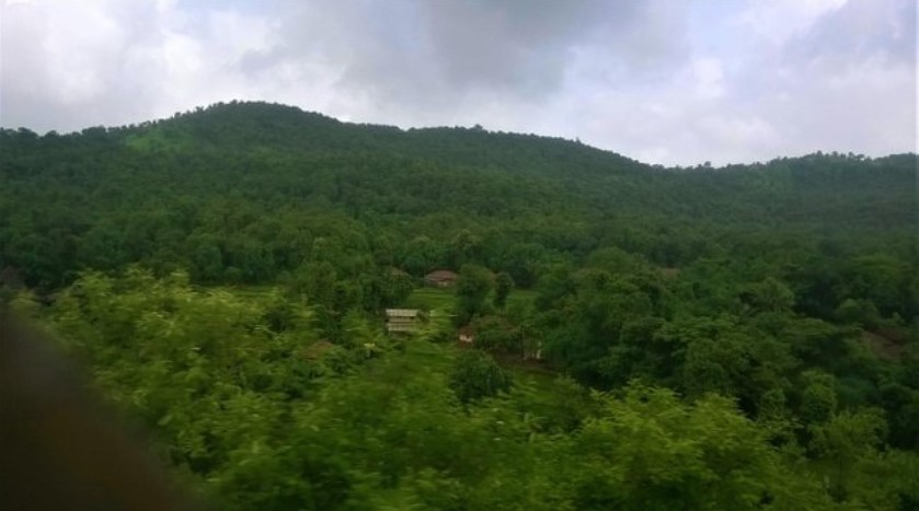 A scenic travel story captured through a train window, showing lush green hills, dense forest, and small village houses nestled in the landscape under a cloudy sky.