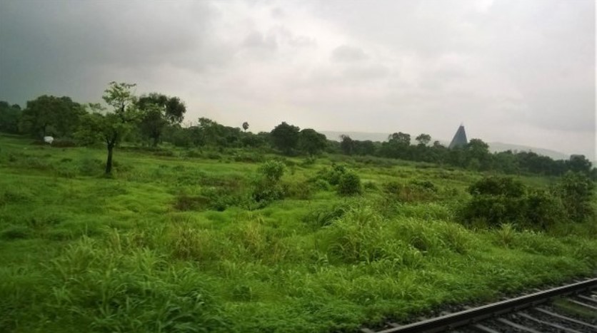 Lush green fields and distant trees seen through a train window, with railway tracks in the foreground and a conical structure on the horizon, capturing a calm monsoon moment in a travel story.