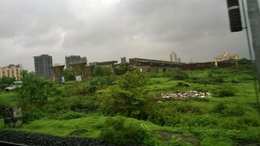 Overgrown green fields with scattered debris, unfinished concrete pillars, and distant city buildings, seen through a train window under a cloudy monsoon sky — a raw and real moment in a travel story.