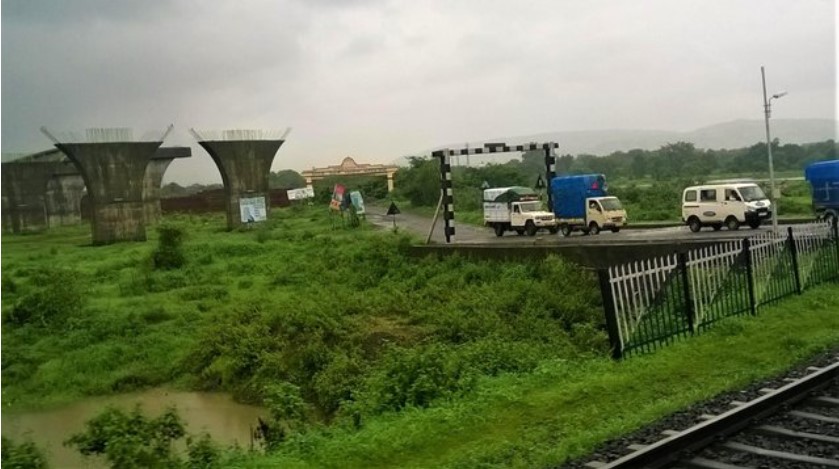 “Partially constructed bridge pillars, a rural level crossing with vehicles, and lush monsoon greenery seen through a train window — a moment that adds contrast and character to a travel story