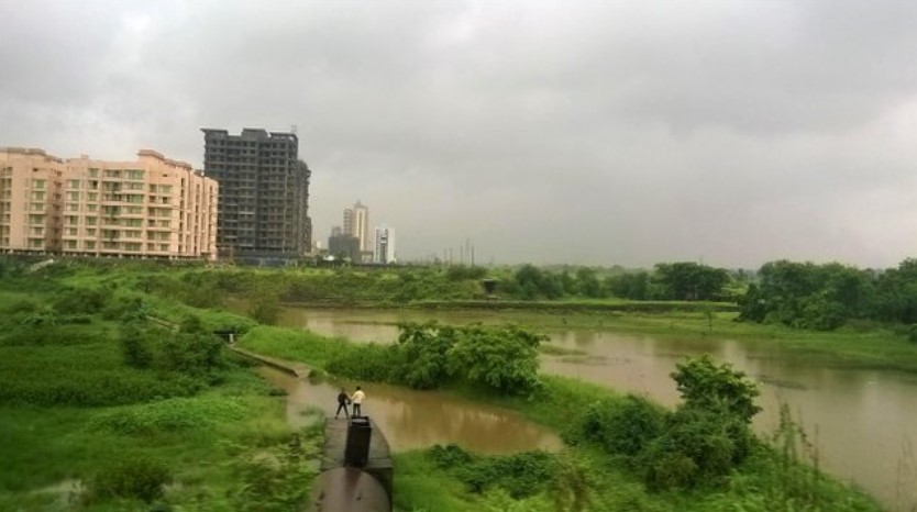 Flooded monsoon fields with two people standing on a small structure, set against tall residential buildings and cloudy skies, seen through a train window — a vivid contrast captured in a travel story.