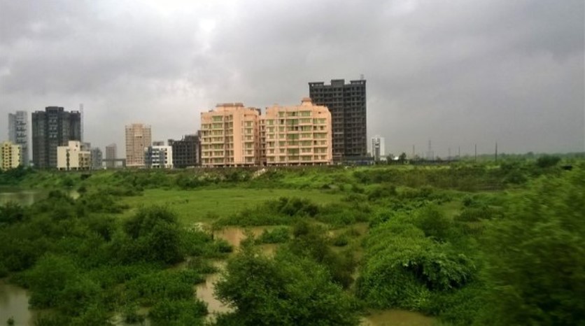 Lush monsoon greenery and flooded patches in the foreground with rising apartment buildings in the distance, seen through a train window — a vivid contrast captured in a travel story