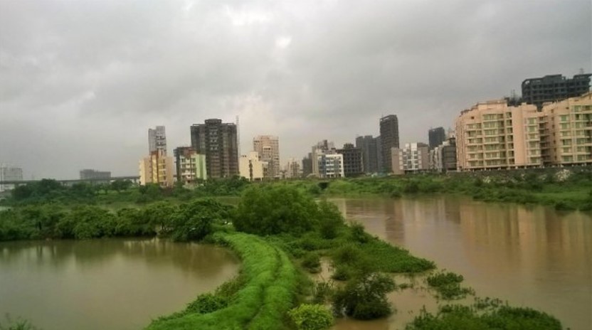 looded monsoon river surrounded by lush greenery with a backdrop of high-rise buildings, viewed through a train window — a striking blend of nature and city in a travel story