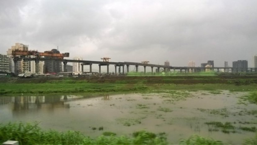 Monsoon-soaked wetlands with a massive elevated bridge under construction and high-rise buildings in the distance, viewed through a train window — capturing a dramatic moment in a travel story