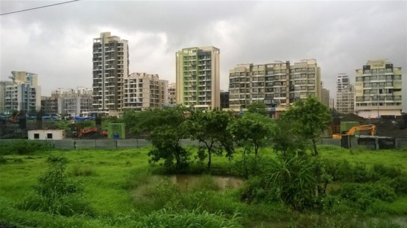 Monsoon-soaked green fields with small water patches and a row of high-rise residential buildings in the background, viewed through a train window — a contrasting scene in a travel story.