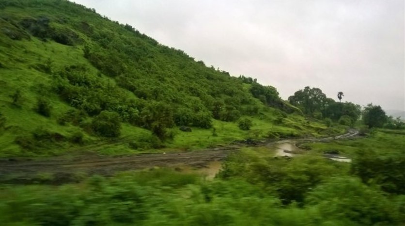 Lush green hillside with a muddy stream winding through the valley, seen through a train window during the monsoon — a refreshing landscape moment in a travel story