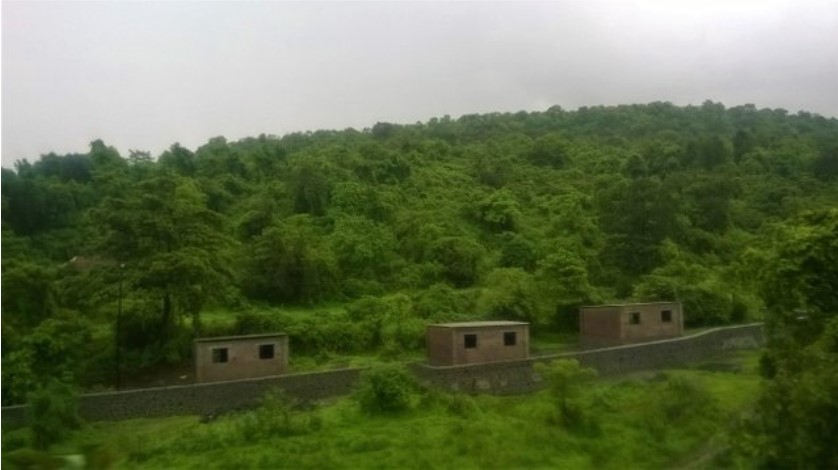 Lush green hills and small brick structures captured through a train window during monsoon, forming a scenic moment perfect for a travel story.