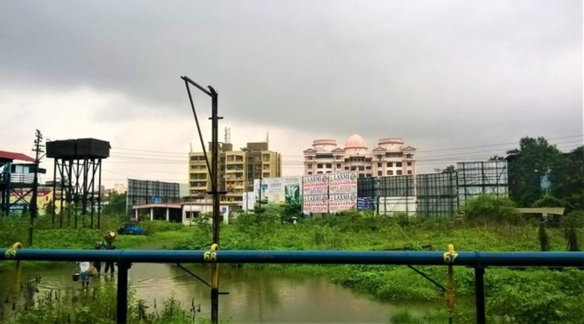 Urban outskirts seen through a train window, with flooded green fields, water tanks, billboards, and residential buildings under cloudy monsoon skies — a contrasting moment in a travel story.