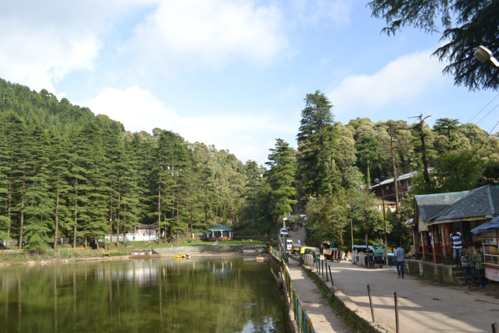 A serene hill-side lake surrounded by dense pine trees, with reflections on the still water, a walkway on the right, small shops, and a few visitors enjoying the peaceful atmosphere.