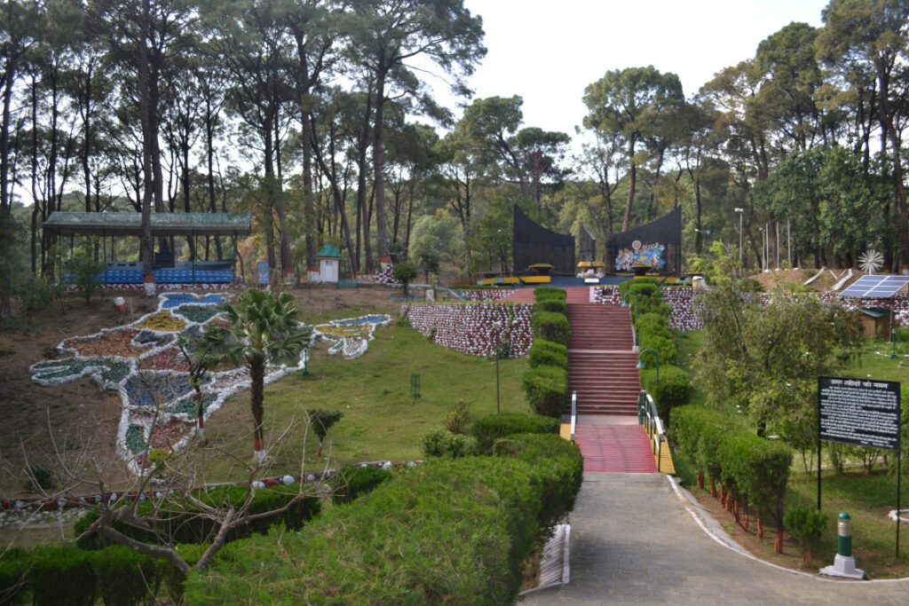 The Dharamshala War Memorial featuring landscaped gardens, a stone map of India, neatly trimmed hedges, red steps leading to the central monument, and tall pine trees forming a serene backdrop.