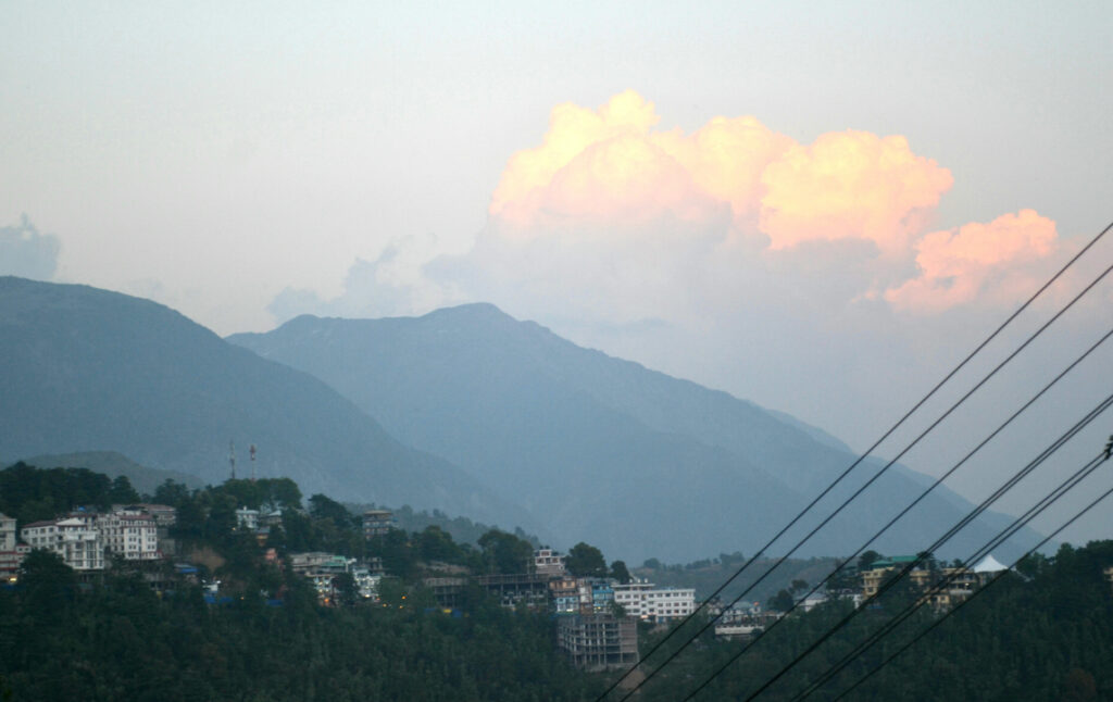 Evening view of a hillside town with layered blue mountains in the background and soft orange clouds glowing above the peaks, with power lines crossing the frame.