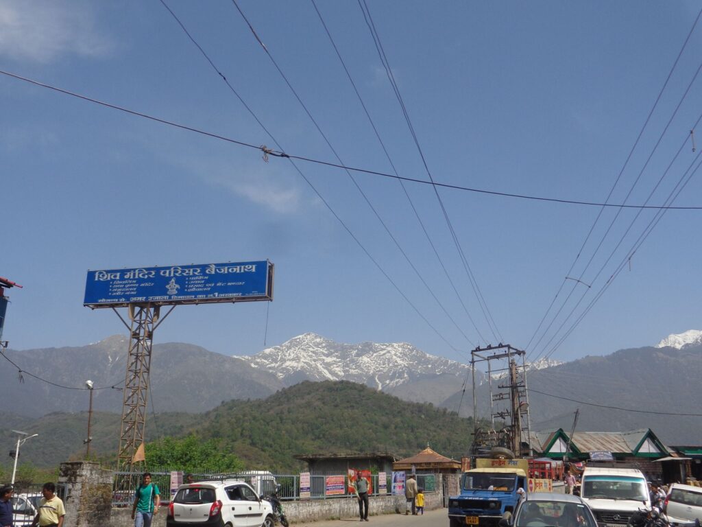 A busy street near Baijnath in dharamshala with vehicles, people, and overhead electric lines, set against a backdrop of snow-capped Himalayan peaks under a clear blue sky.