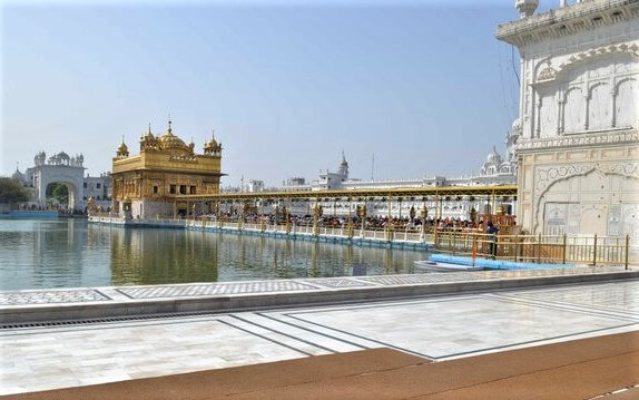 Golden Temple in Amritsar reflecting in the calm waters of the Amrit Sarovar, with the marble walkway and surrounding white structures under a clear blue sky.