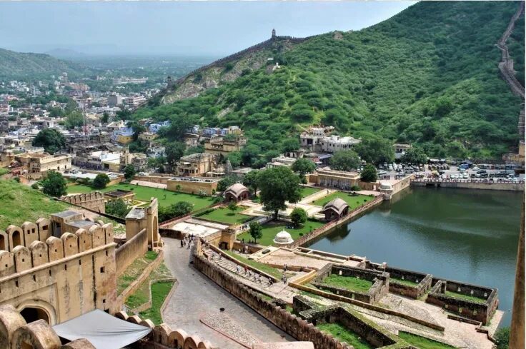A panoramic view of Amer Fort in Jaipur, India, showing its historic ramparts, gardens, and the surrounding Aravalli hills with Maota Lake beside it.