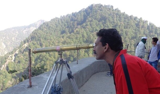A man in a red jacket observing the distant mountains through a brass telescope at the Himalayan View Point, surrounded by forested hills.