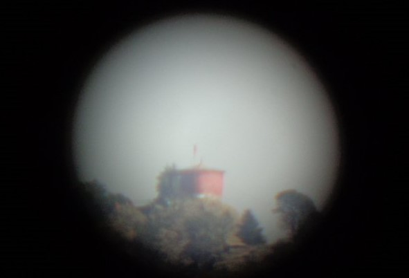 A distant red temple structure on a hilltop surrounded by trees, viewed through a telescope’s circular lens at the Himalayan View Point.