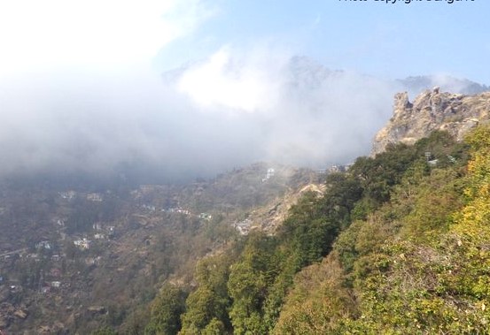 Mist-covered mountains with scattered greenery and rocky outcrops under a partly cloudy sky. view from patni top in nainital
