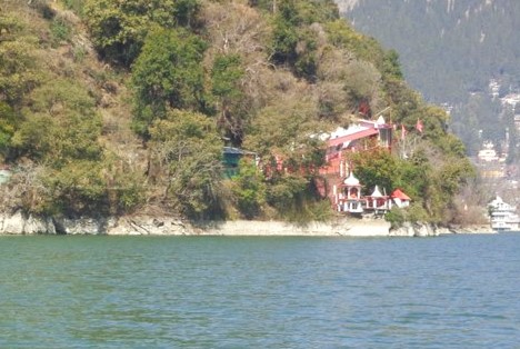 A lakeside temple complex surrounded by trees and hills in Nainital, with calm blue water in the foreground.