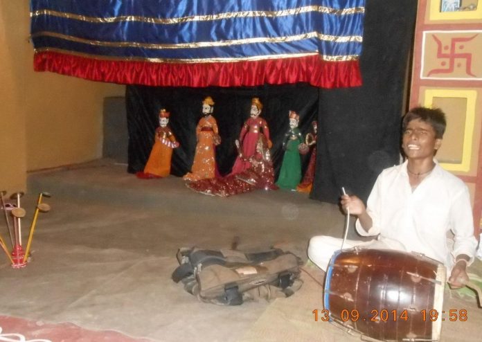 A traditional Rajasthani puppeteer performing with colorful string puppets on stage, accompanied by live drum music at Chokhi Dhani in Jaipur.
