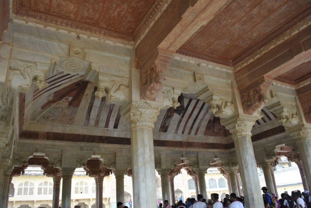 Intricately carved sandstone arches and pillars inside the Diwan-i-Aam (Hall of Public Audience) at Amber Fort, Jaipur, showcasing beautiful Mughal-Rajput architectural details and a gathering of visitors beneath the ornate ceiling.