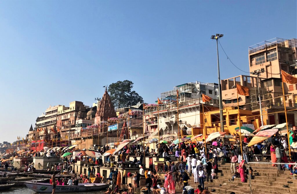 A lively morning scene at Dashashwamedh Ghat in Varanasi, with pilgrims, colorful umbrellas, and temple spires along the banks of the Ganges under a clear blue sky.