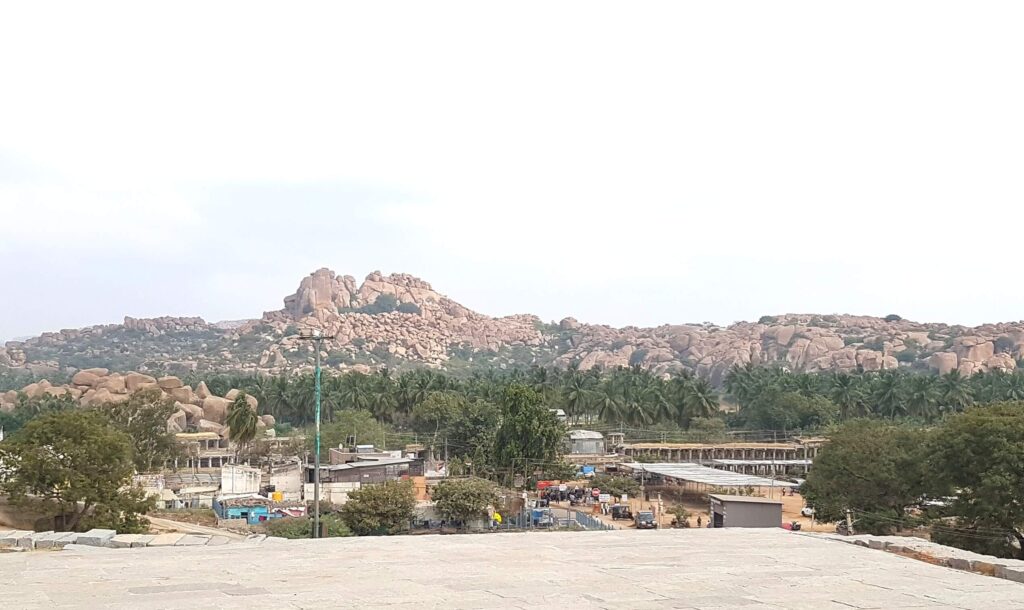 A panoramic view of Hampi’s rocky hills scattered with massive boulders, coconut trees lining the foreground, and small village structures beneath a cloudy sky.
