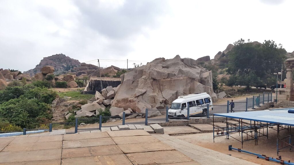 A rocky landscape in Hampi with giant boulders, stone steps, and a white tourist van parked beside ancient ruins under a cloudy sky.
