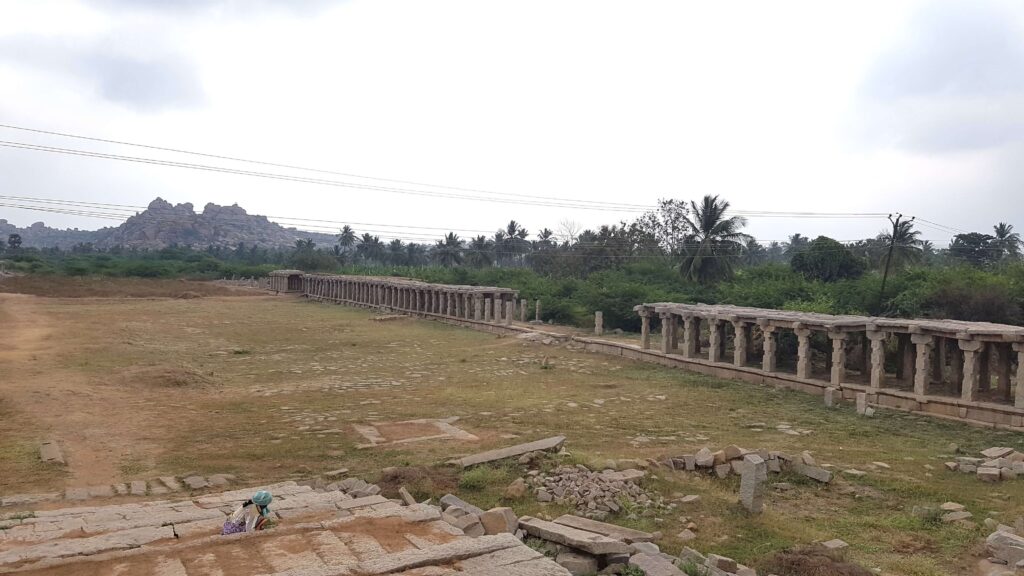 A long row of ancient stone pillars stretching across an open field in Hampi, with rocky hills and palm trees in the distance.