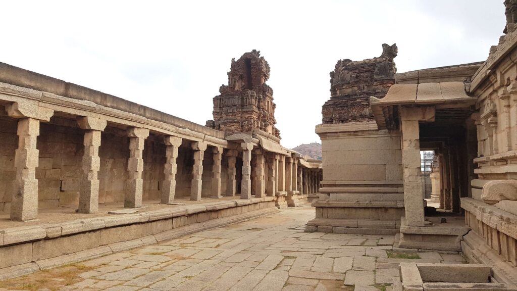 A long stone corridor with carved pillars and partially ruined temple towers at Hampi, showcasing the grandeur of ancient architecture.