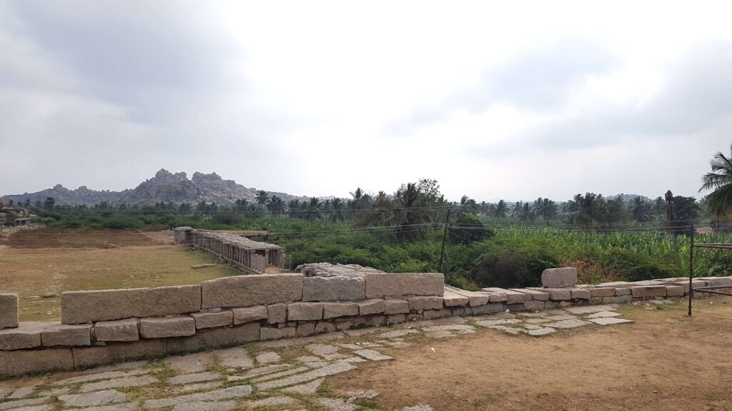 A serene view of the Hampi landscape with ancient stone ruins in the foreground, lush greenery, and rocky hills in the distance under a cloudy sky.