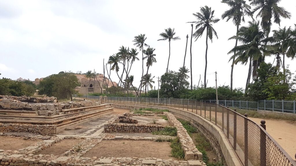 Ancient stone ruins in Hampi surrounded by tall palm trees, with rocky hills and an old structure visible in the background under a cloudy sky.