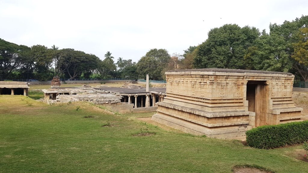 Ancient stone structures of Hampi surrounded by green lawns and trees, showing remnants of temples and pillared halls.