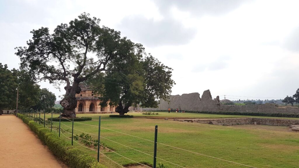Ancient palace ruins in Hampi surrounded by green lawns, old trees, and remains of a stone fort wall.