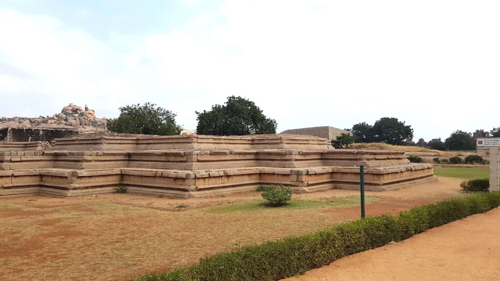 Ancient stone platform ruins in Hampi, surrounded by dry earth and greenery, with rocky hills visible in the background.