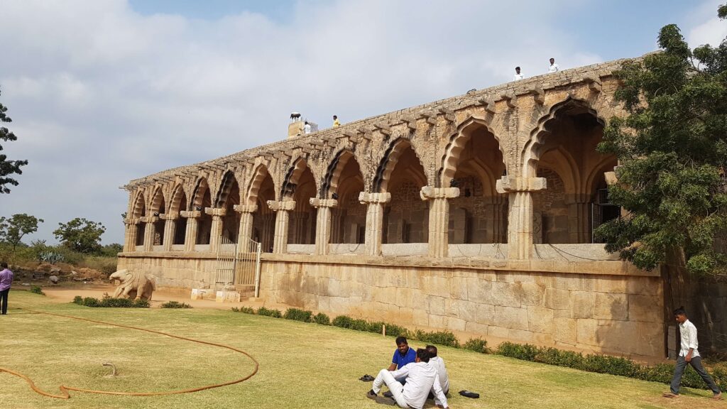 A long stone structure with a row of arched domes and ornate pillars, known as the Elephant Stables in Hampi, with visitors sitting and walking in the grassy courtyard.
