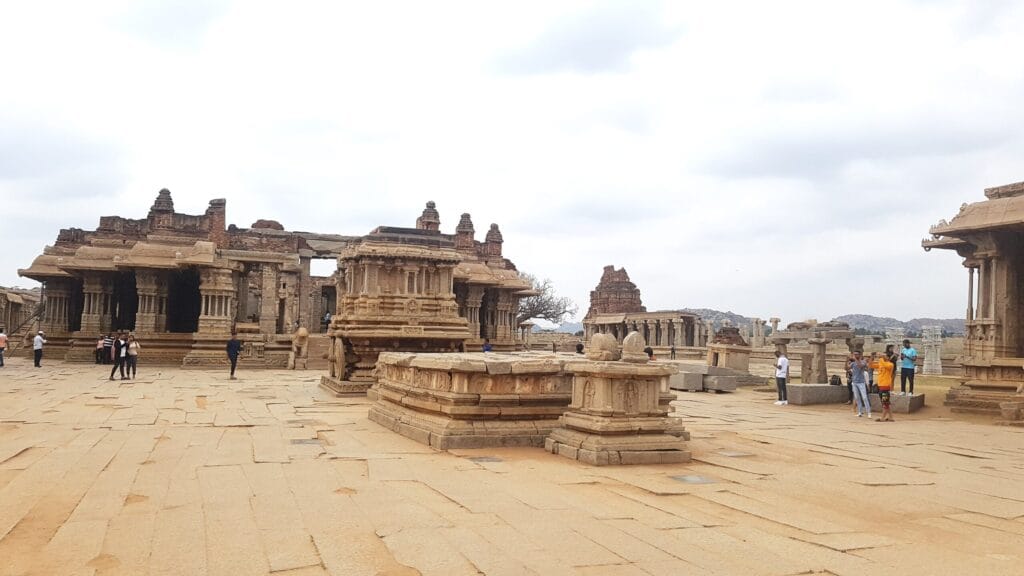 Ancient stone structures and the iconic stone chariot at Hampi’s Vittala Temple complex, with visitors walking across the vast courtyard under a cloudy sky.