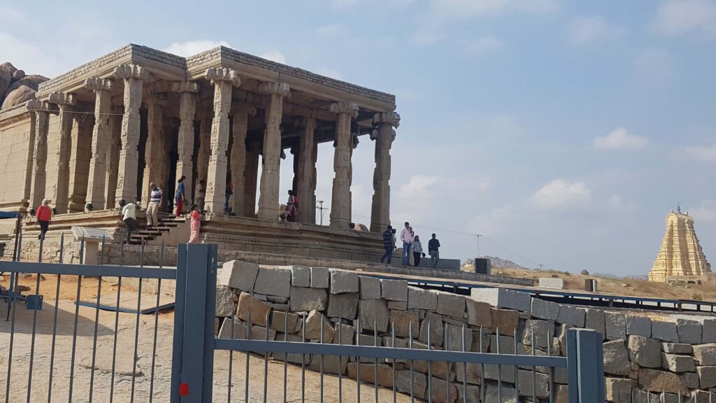 Stone mandapa with intricately carved pillars at Hampi, with the Virupaksha Temple tower visible in the distance.
