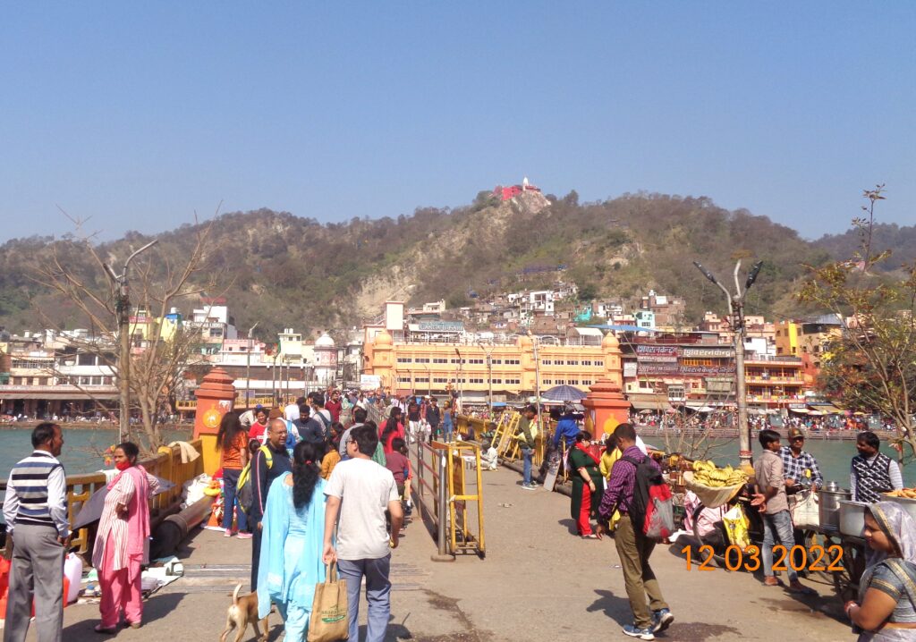 Pilgrims and visitors walking across the bridge at Haridwar with colorful ghats and hills and mansa devi temple at hill top in the background
