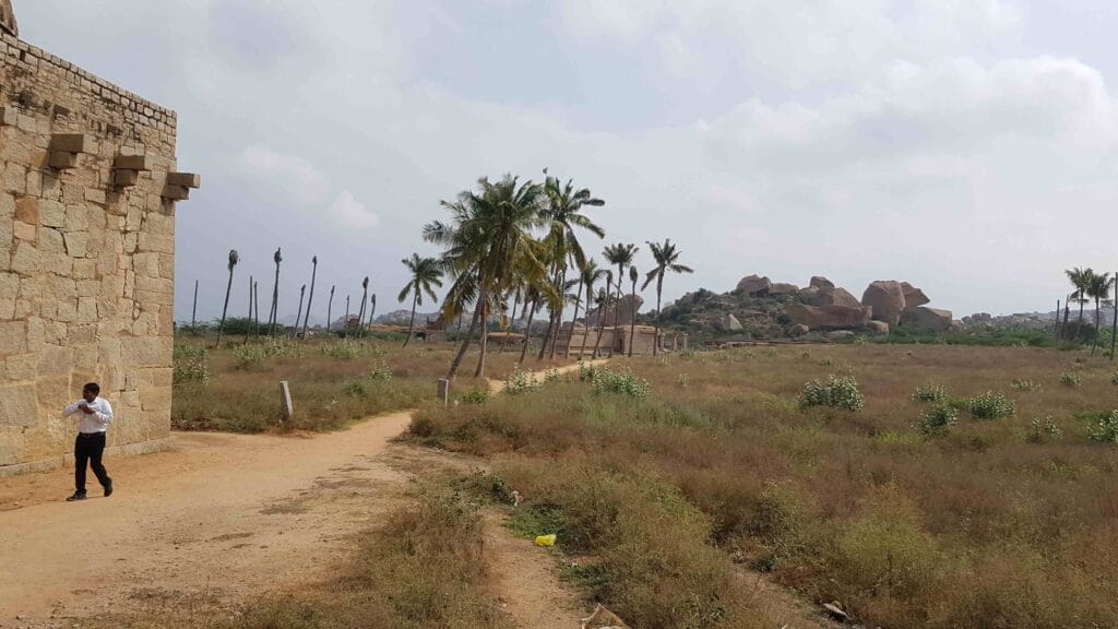A quiet pathway beside an old stone wall in Hampi, leading through dry grasslands with tilted palm trees and large rocky boulders scattered across the distant hillside.