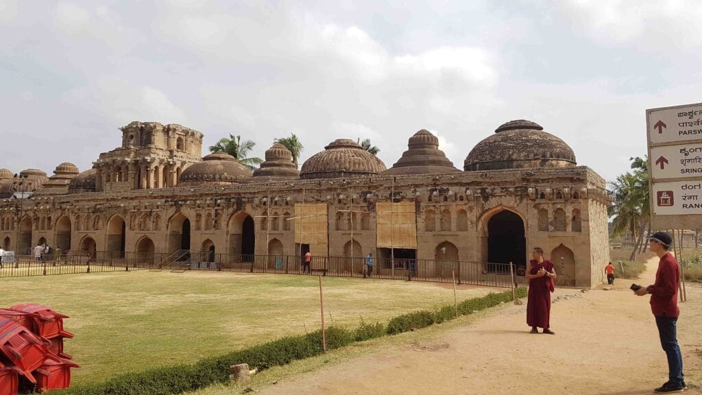 The Elephants’ Stables at Hampi, featuring a long row of arched chambers topped with distinct domes in Indo-Islamic style, with visitors walking around the well-kept lawn in front.