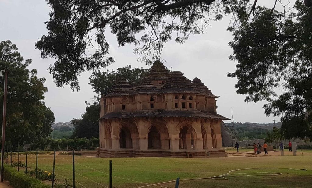 The Lotus Mahal in Hampi, a two-storey pavilion with elegant arches and a tiered pyramidal roof, surrounded by greenery and visitors walking around the lawn.