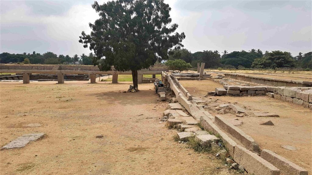 Ancient stone aqueduct remains at Hampi, with long aligned stone channels, scattered blocks, and a large tree at the center of the open archaeological site.
