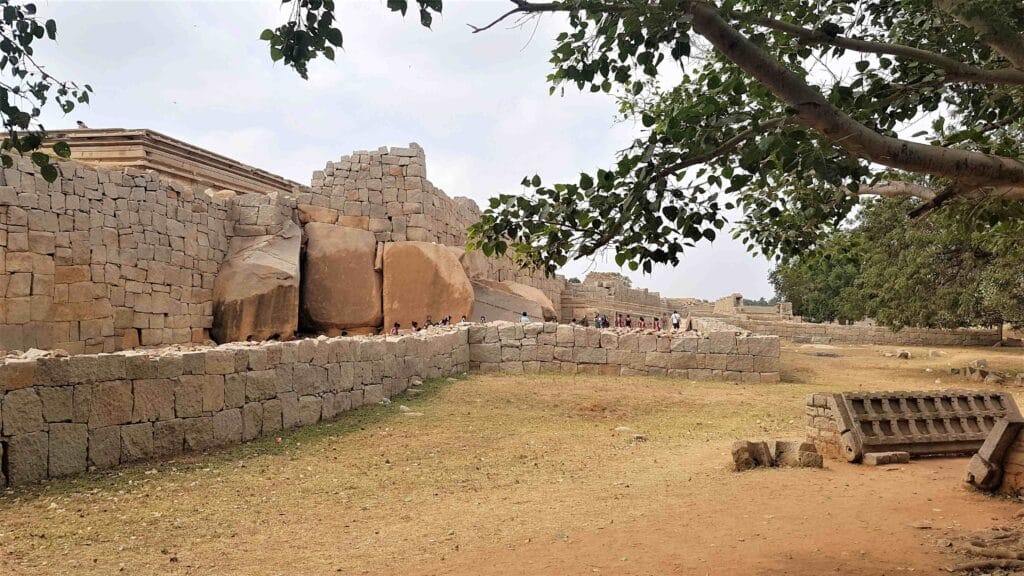 A section of the ruined fortification walls at Hampi, featuring large boulders integrated into stone masonry, with visitors walking along the pathway and trees framing the scene.