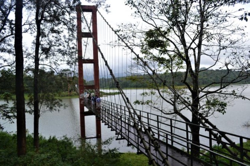 A suspension bridge with red support towers spans across a calm lake, surrounded by trees and lush greenery. A group of people is seen walking along the narrow bridge, with forested hills and a cloudy sky in the background.