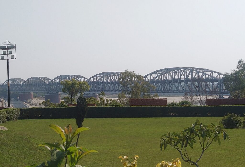 View of the Malviya Bridge in Varanasi, a double-decker steel truss bridge stretching across the River Ganga, seen from a lush green riverside garden under a clear sky.